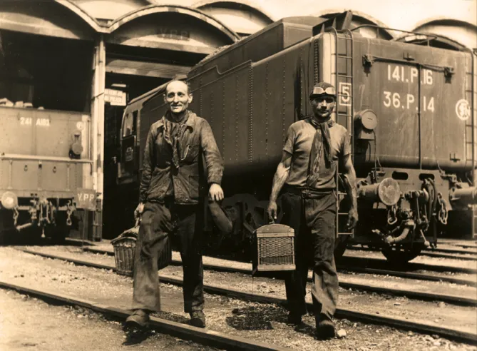 Ein Mechaniker (rechts) und sein Fahrer verlassen in den 1960er Jahren ihr Depot. Sie beenden einen harten Tag an Bord ihrer 141-P und nehmen ihren traditionellen Korb (daher der Ausdruck „Lunchpaket“) mit, der die Reise in den hinteren Kästen des Tenders angetreten hat.© IXO Collections SAS - Tous droits réservés. Crédits photo ©&nbsp;Collection Pierre Decroix.