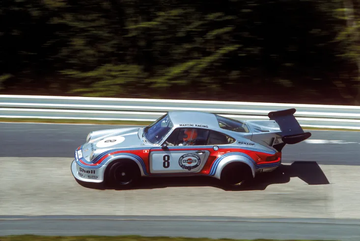 Ein Porsche 911 RSR Turbo 2.1 zeigt während einer Vorführung im englischen Goodwood seinen großen mattschwarzen Flügel, der auf Stützhalterungen montiert ist, die direkt an den hinteren Pfosten befestigt sind.© IXO Collections SAS - Tous droits réservés. 